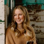 Blonde woman in beige jumper smiling looking out of window holding her smart phone