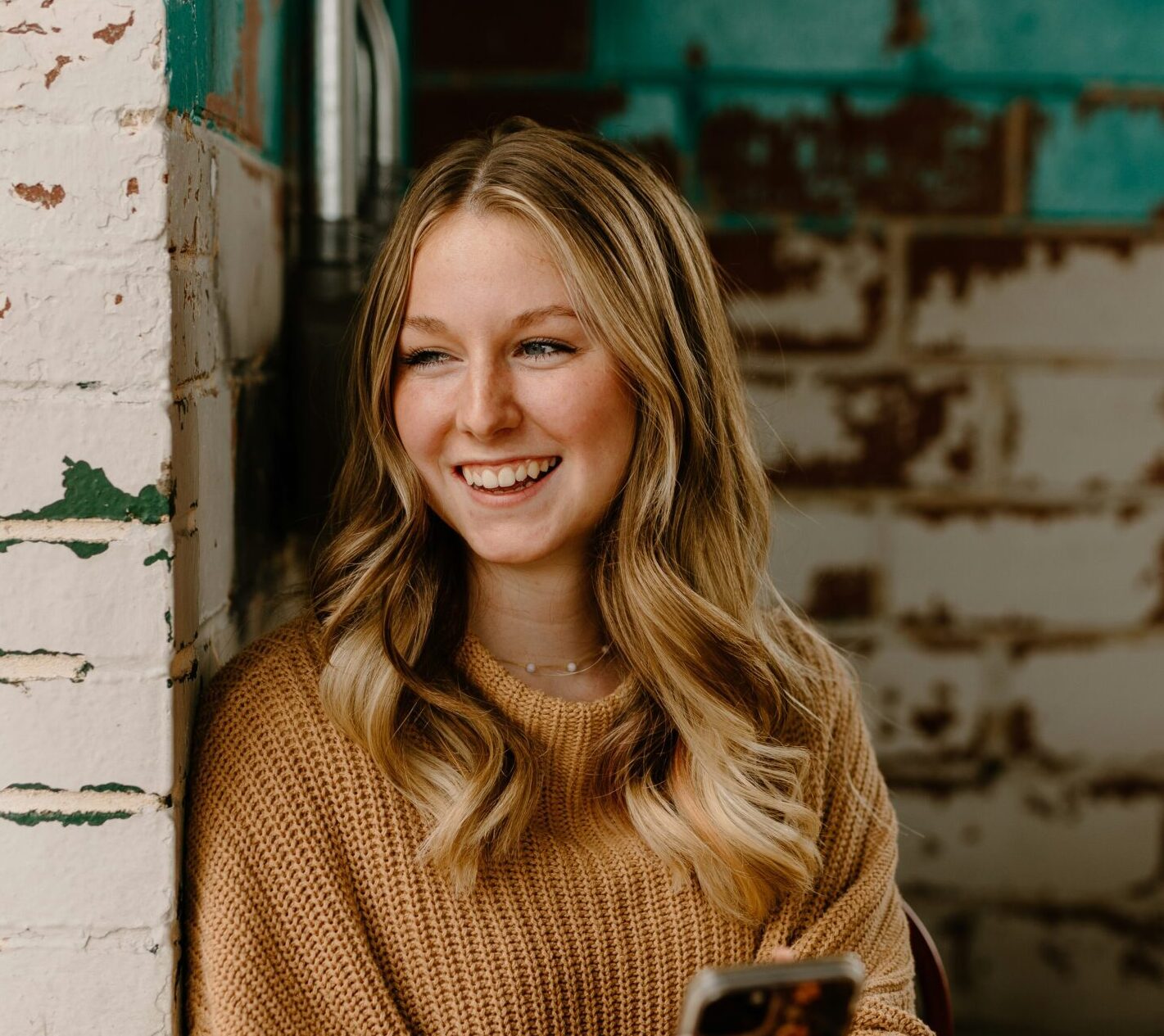 Blonde woman in beige jumper smiling looking out of window holding her smart phone