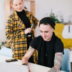 Woman in yellow checkered jacket with her hair in a bun and a man with tattoos wearing a black t-shirt looking at a laptop