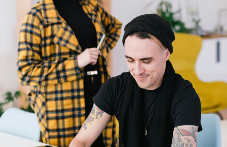 Woman in yellow checkered jacket with her hair in a bun and a man with tattoos wearing a black t-shirt looking at a laptop