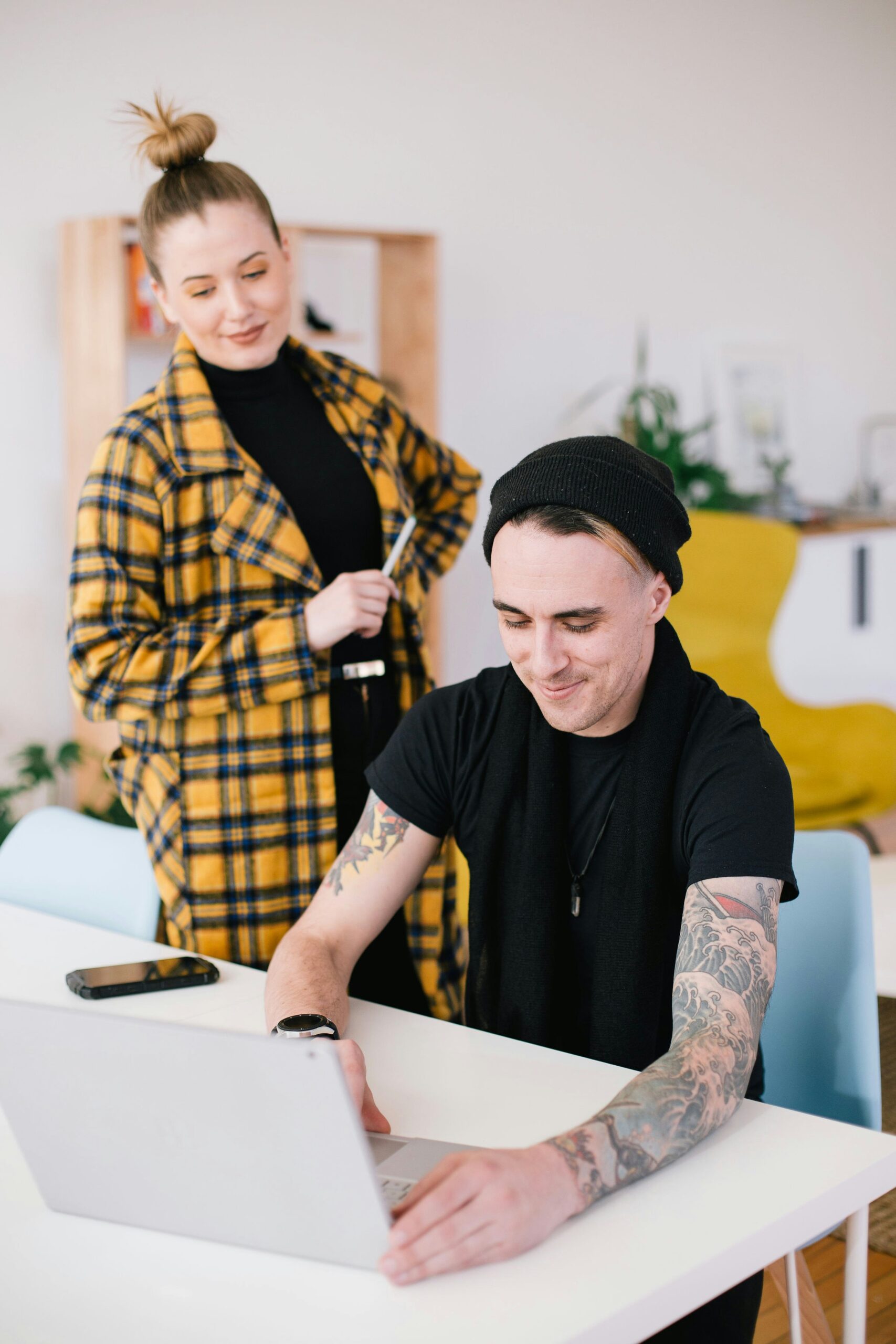 Woman in yellow checkered jacket with her hair in a bun and a man with tattoos wearing a black t-shirt looking at a laptop