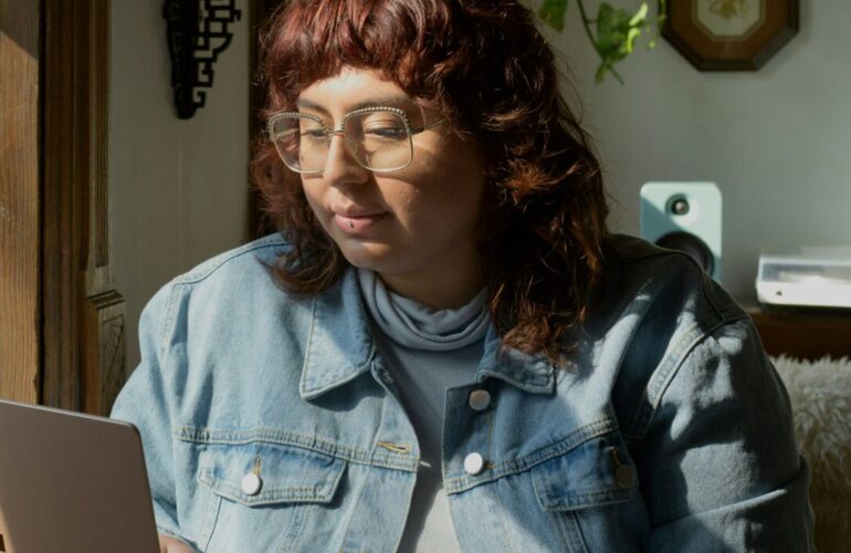 woman in blue denim jacket and glasses working at a laptop in sunlight
