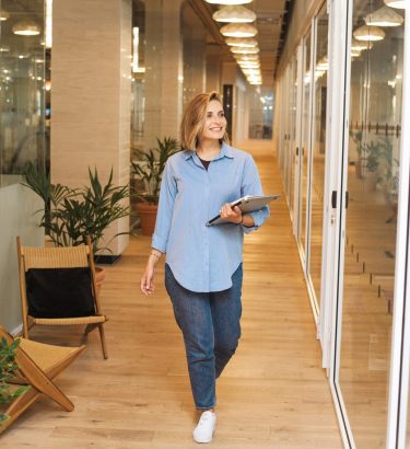 lady-blue-denim-office-walking lady in blue denim smiling walking in an office holding a laptop